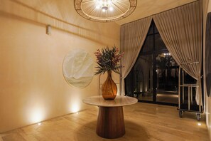 A round wooden table with a vase of flowers, a wall-mounted light fixture, and a view of the outdoors through a large window with curtains.