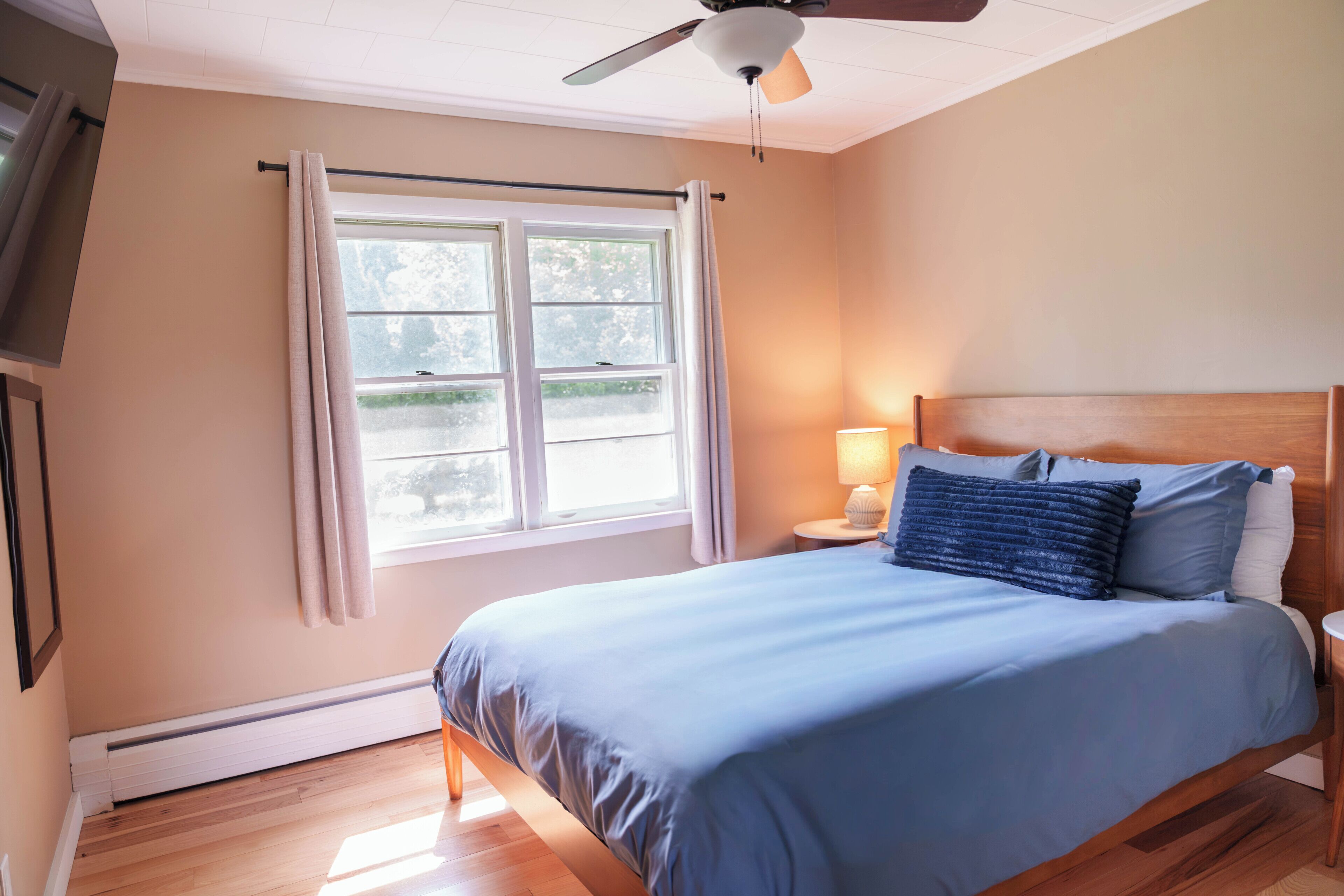 A bedroom with a bed, a nightstand, a lamp, a ceiling fan, and a window with curtains.
