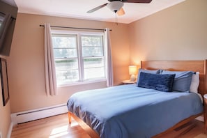 A bedroom with a bed, a nightstand, a lamp, a ceiling fan, and a window with curtains. - The Rutland Ranch: 3 BR/1BA Renovated Home Close to Everything (Rutland)