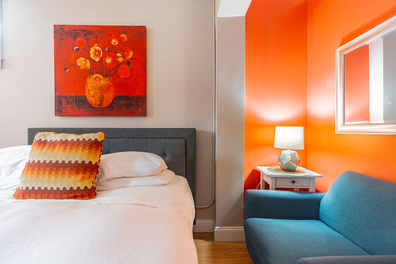 Habitación de hotel con cama, un sillón azul, una mesita de noche con lámpara y un cuadro en la pared.
