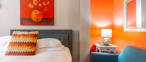 Habitación de hotel con cama, un sillón azul, una mesita de noche con lámpara y un cuadro en la pared.