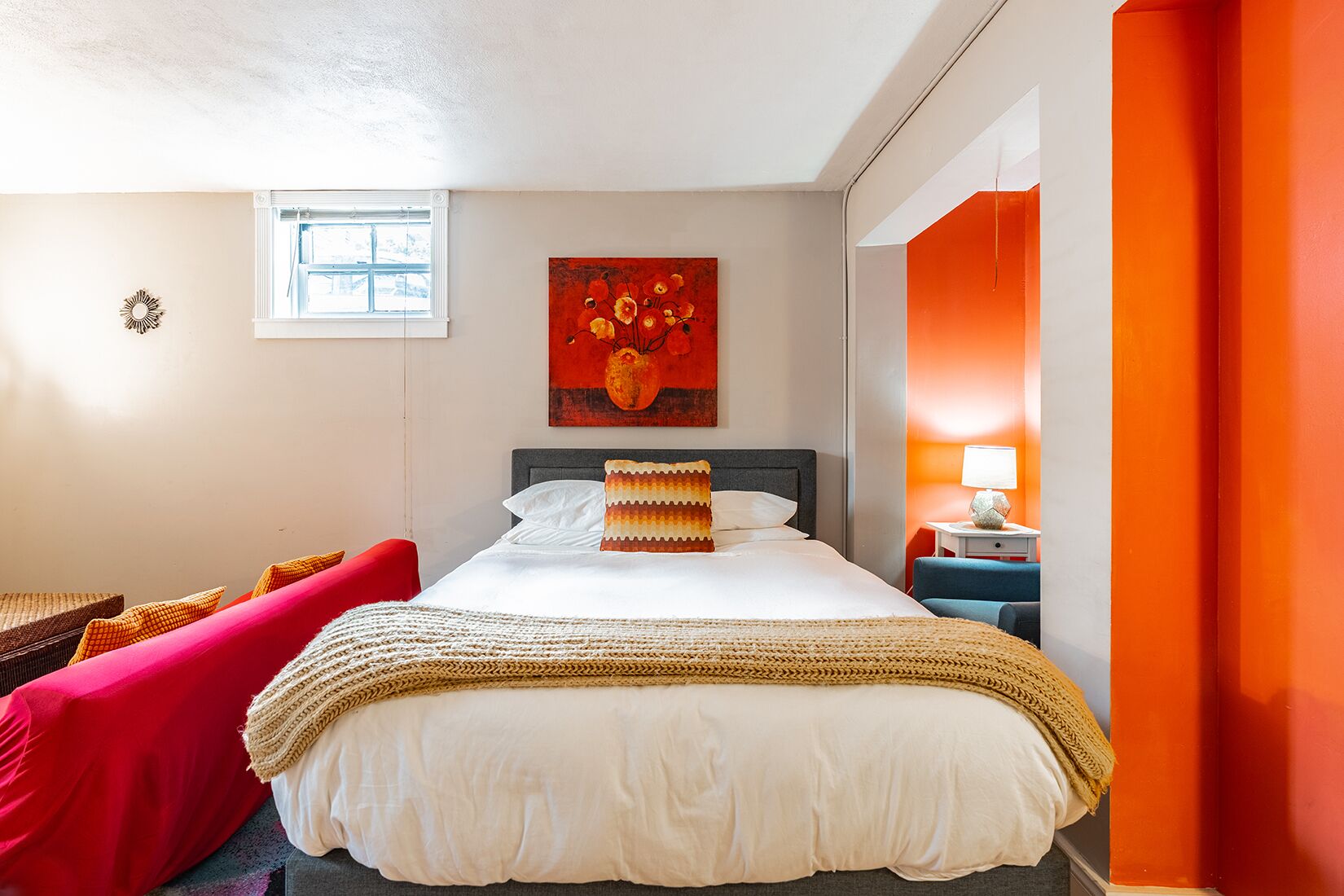 Une chambre d’hôtel avec un lit, un fauteuil rouge, une petite table avec une lampe et un tableau au mur.