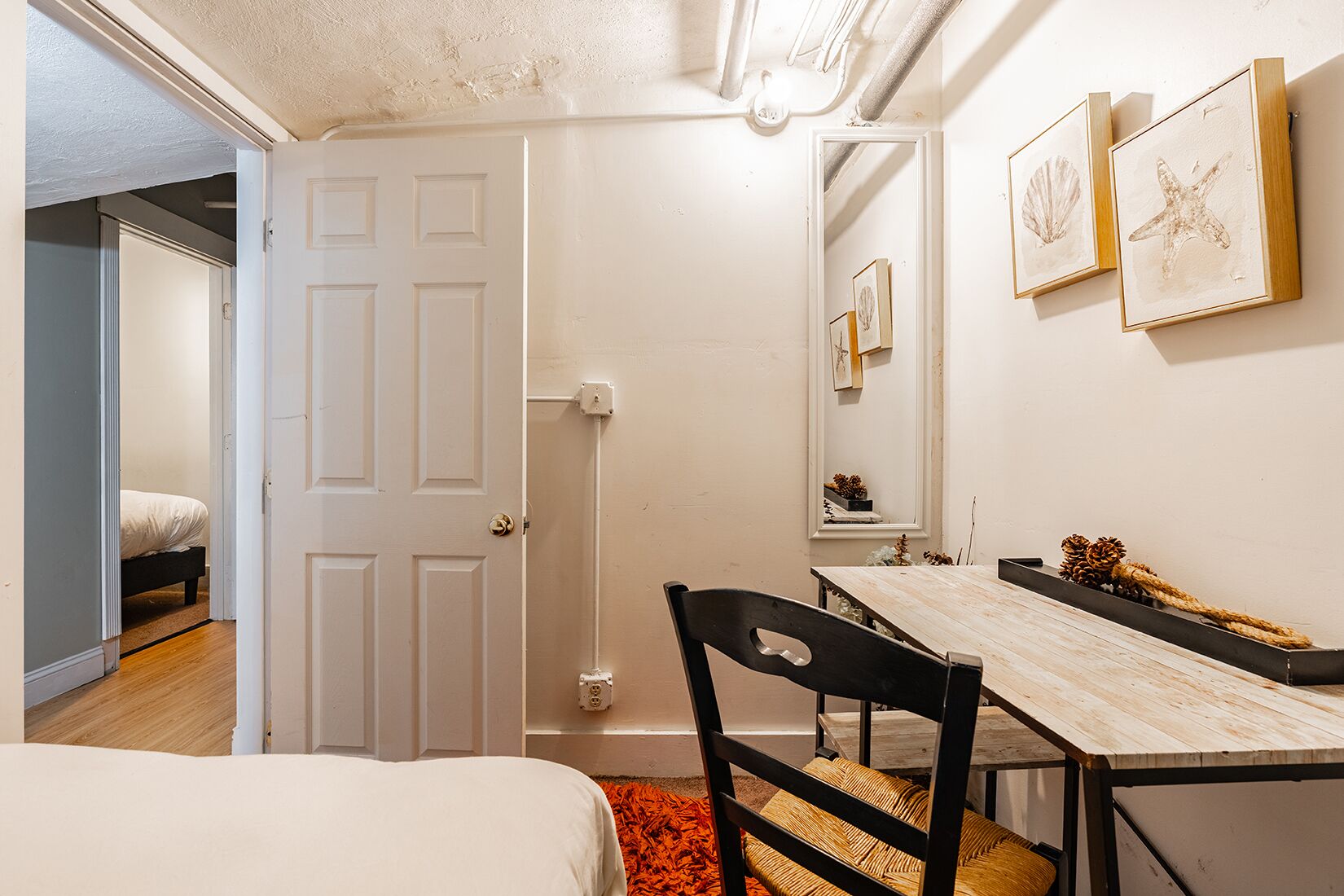 Un dormitorio con una cama, un escritorio de madera y una silla. Hay una puerta que conduce a otra habitación y un espejo en la pared.