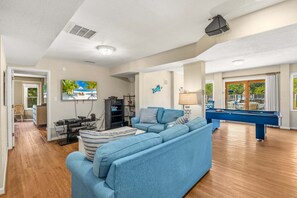 A spacious living room with a blue sofa, a pool table, and a television mounted on the wall. - Family Affair Corolla retreat with private pool, hot tub, and easy beach access. (Corolla)