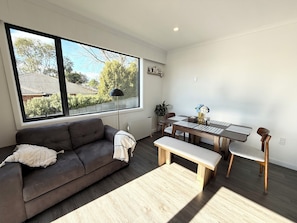 A modern living room with a sofa, a dining table with chairs, a bench, and a large window with a view of trees and a house.