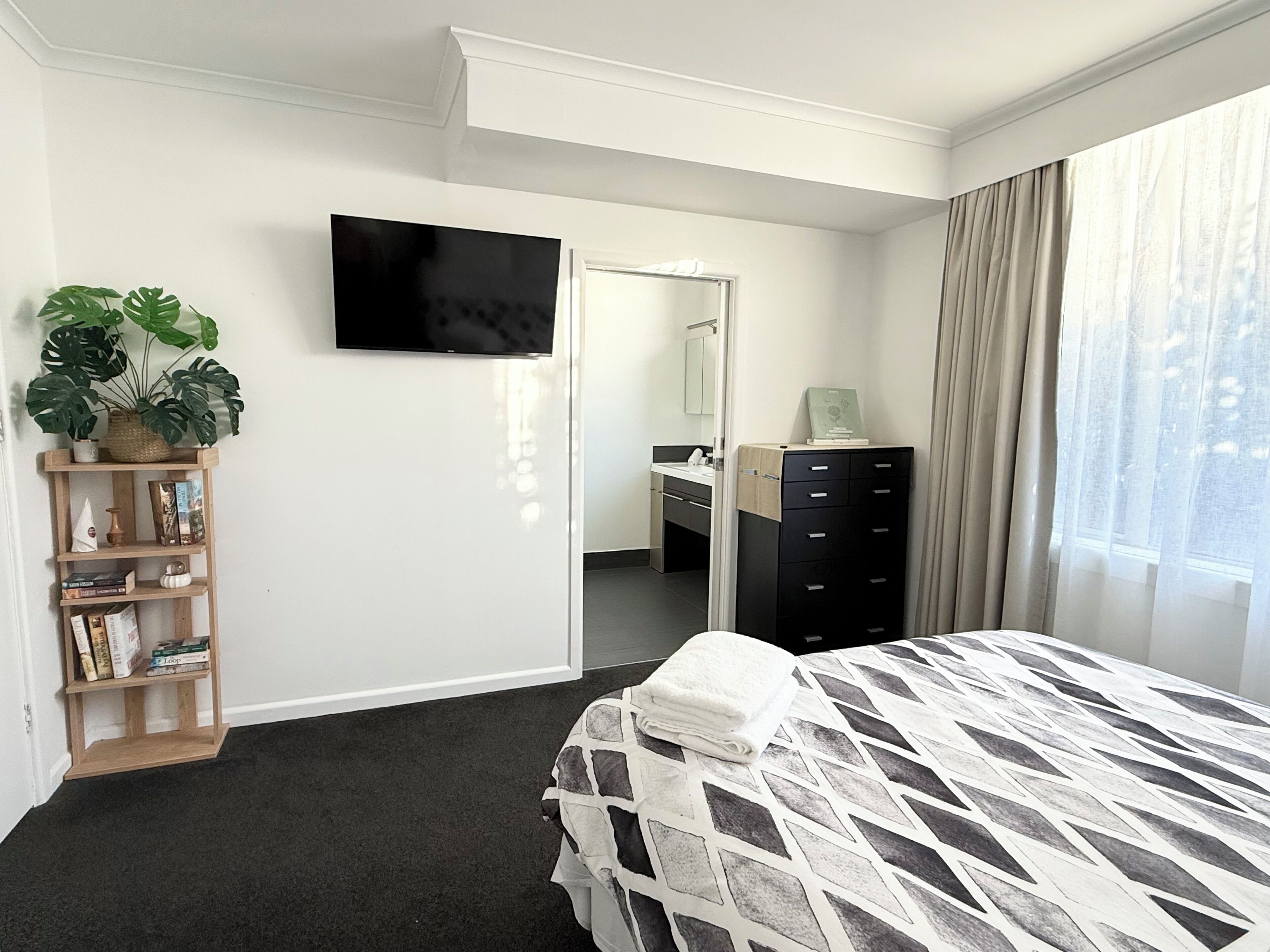 A hotel room with a bed, a television, a dresser, a mirror, a plant, and a bookshelf.