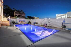 Une piscine rectangulaire à l’eau légèrement bleutée, entourée d’une terrasse avec des chaises longues et une pancarte sur laquelle on peut lire : « Bienvenue au paradis de la plage ».