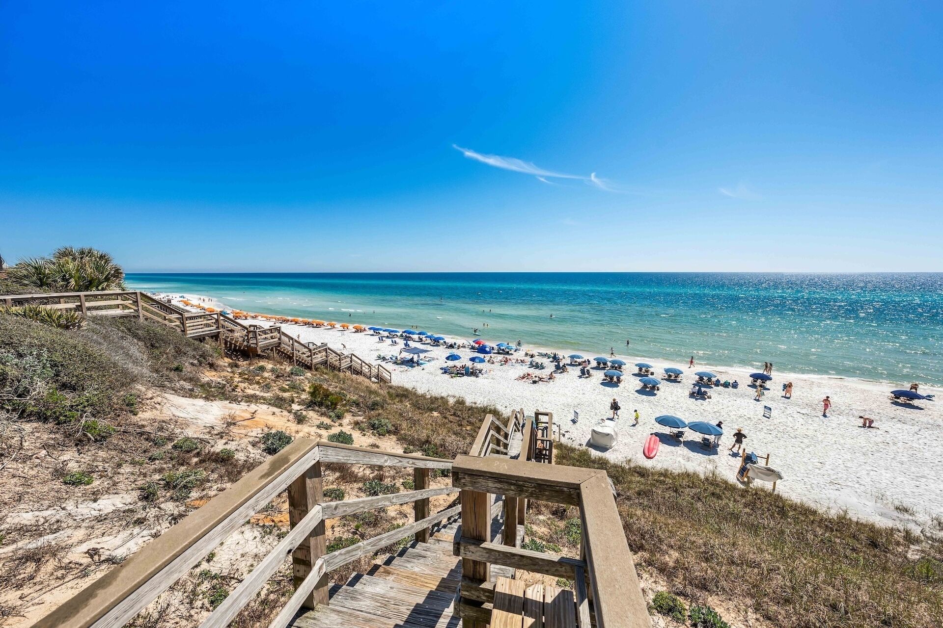 A beach with a wooden boardwalk leading to the shore, clear blue water, and beach umbrellas.