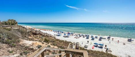 A beach with a wooden boardwalk leading to the shore, clear blue water, and beach umbrellas.