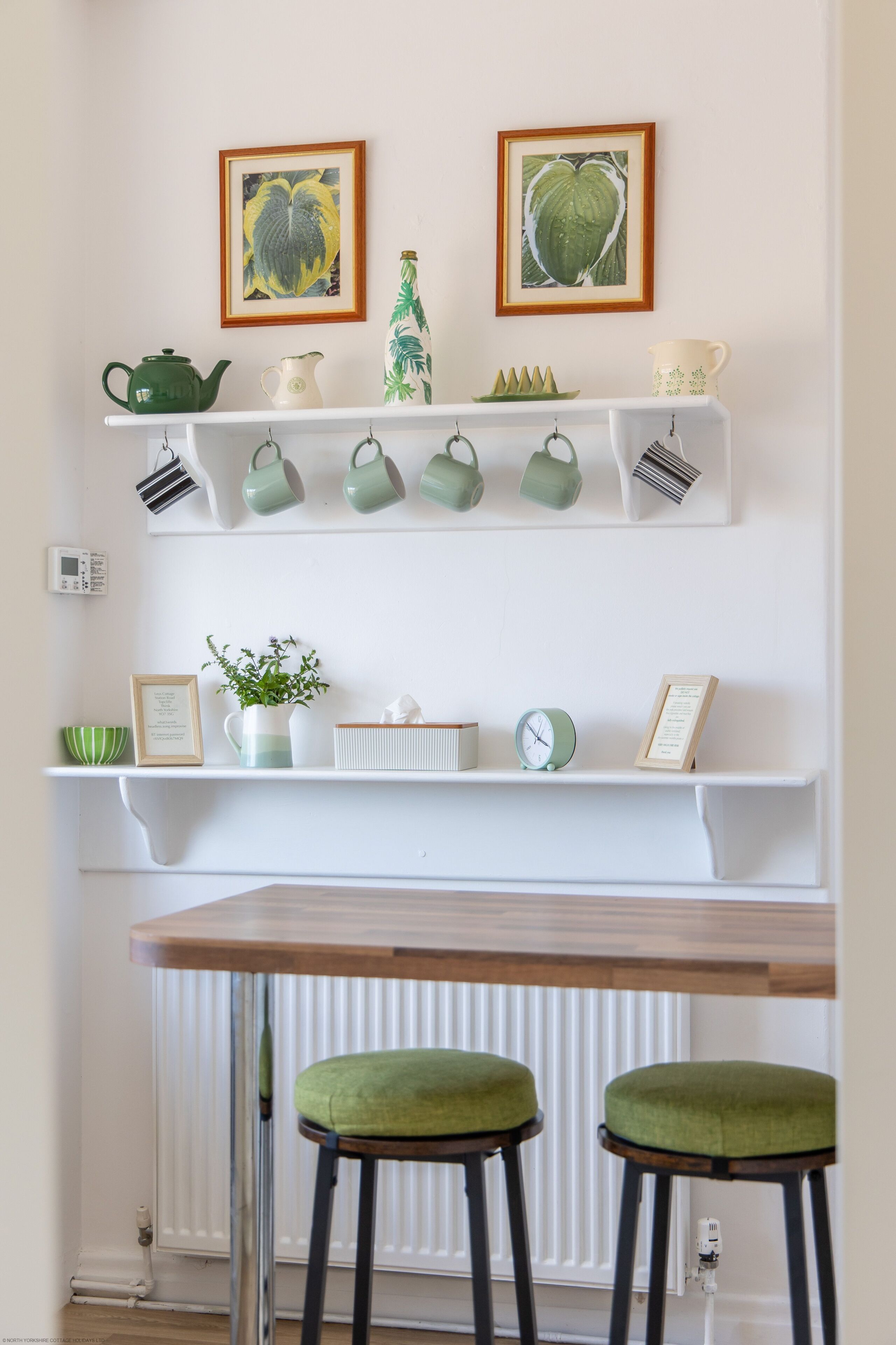 A kitchen area with two shelves holding kitchenware, a small plant, and a framed picture. Below the shelves, there is a wooden table with two green stools.