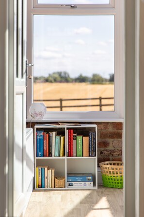 A bookshelf with books and a jigsaw puzzle box, a wicker basket, and a view of a brick wall and open field through a window. - Leys Cottage - sleeps 2 guests (TOPCLIFFE)