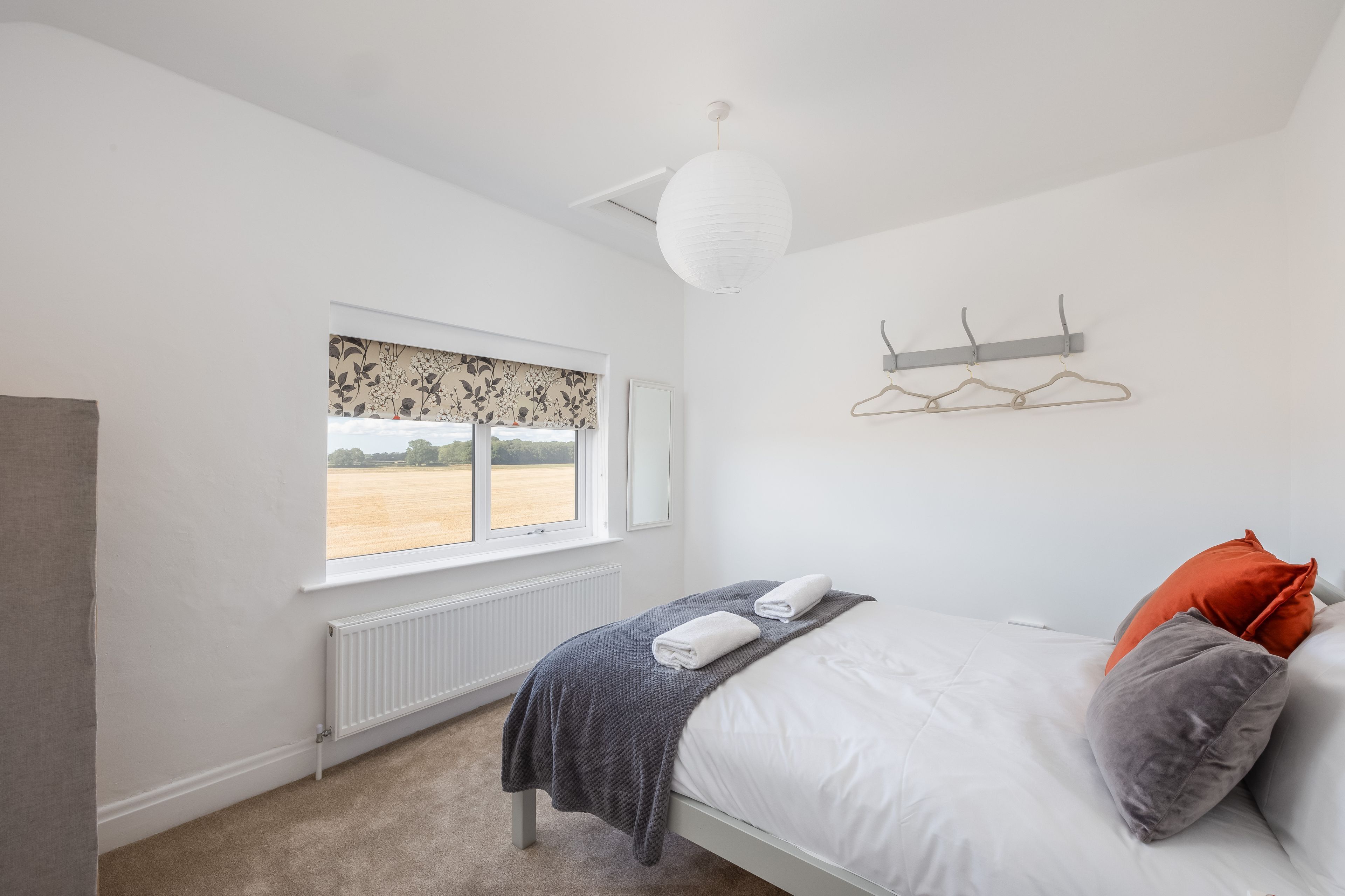 A bedroom with a bed, a radiator, a window with a view of a field, and a hanging light fixture.