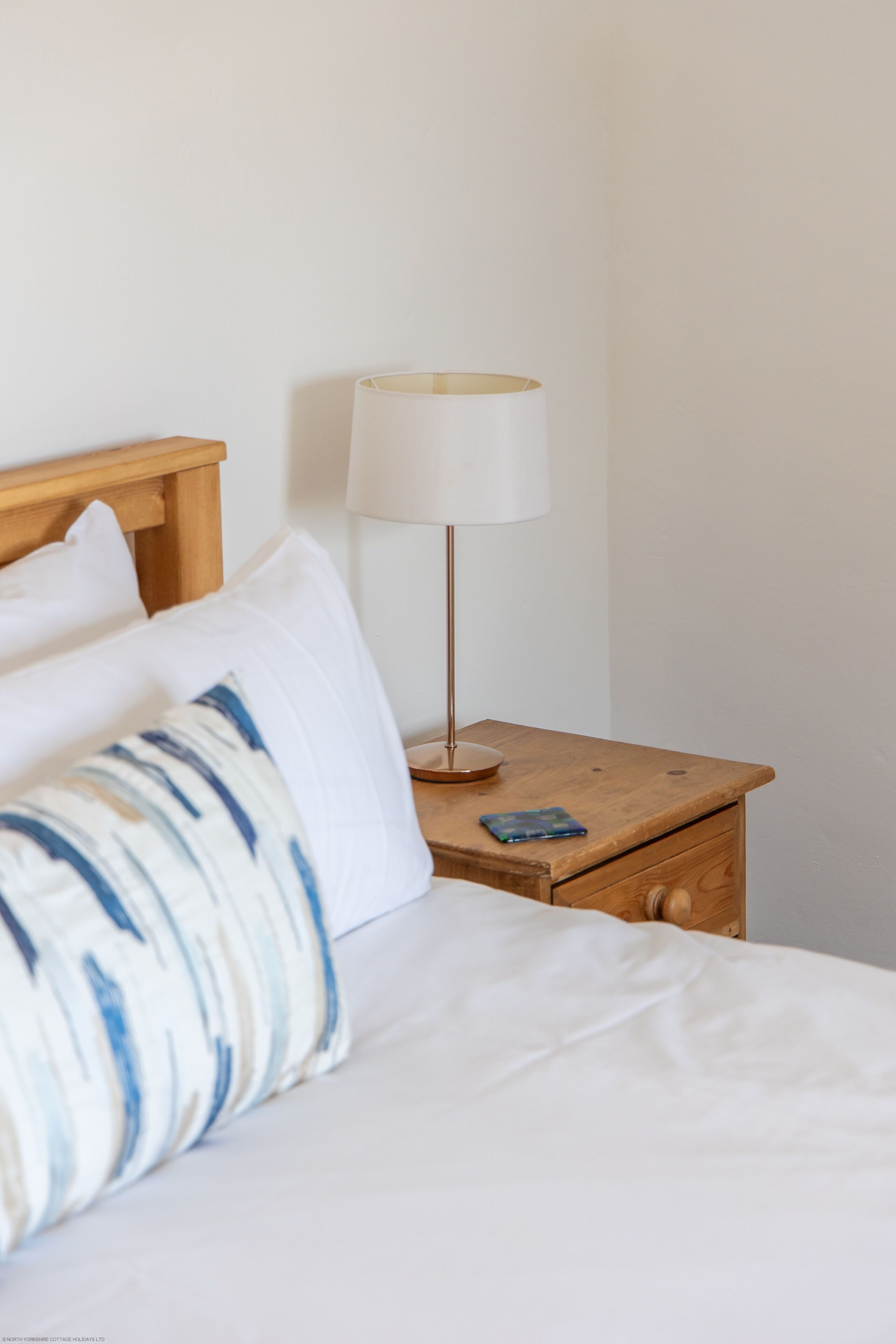 Un lit bien fait, avec des oreillers blancs et bleus à motifs, une table de chevet en bois sur laquelle se trouve une lampe blanche, et un petit tiroir.