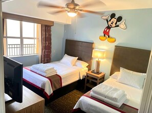 Ett hotellrum med två sängar, en takfläkt, en tv och en väggdekor med Musse Pigg.