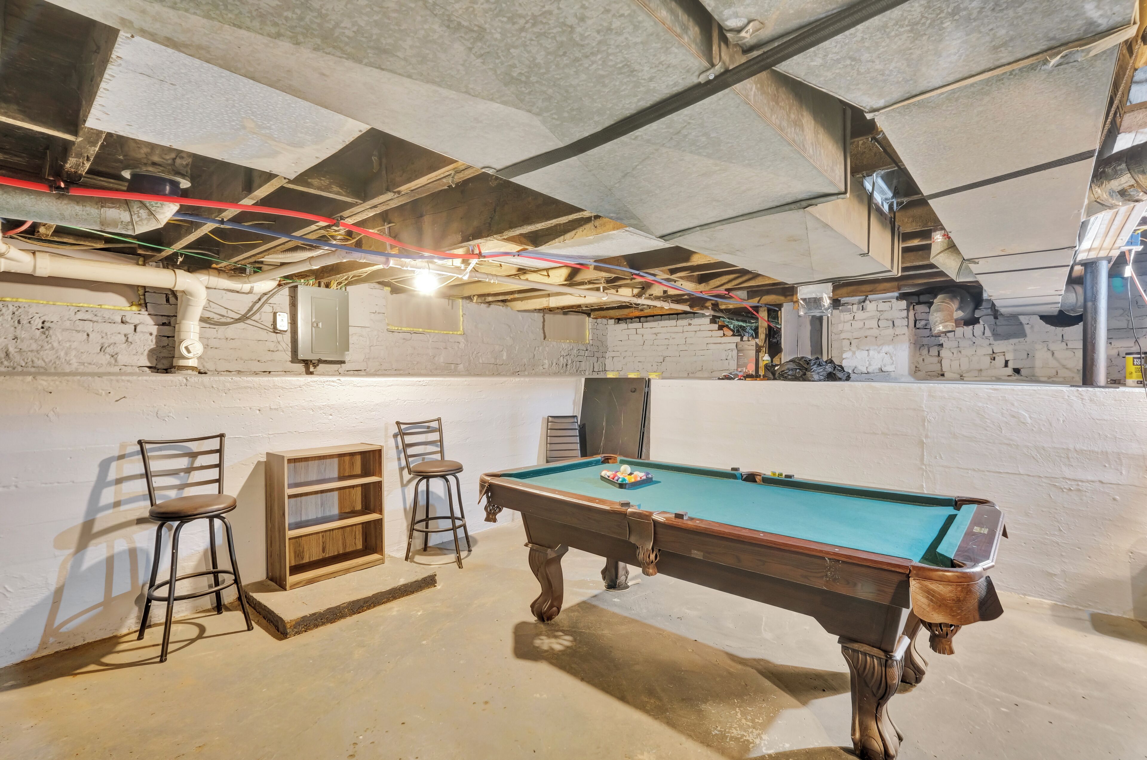 A pool table in a room with a brick wall, two bar stools, and exposed ceiling pipes.