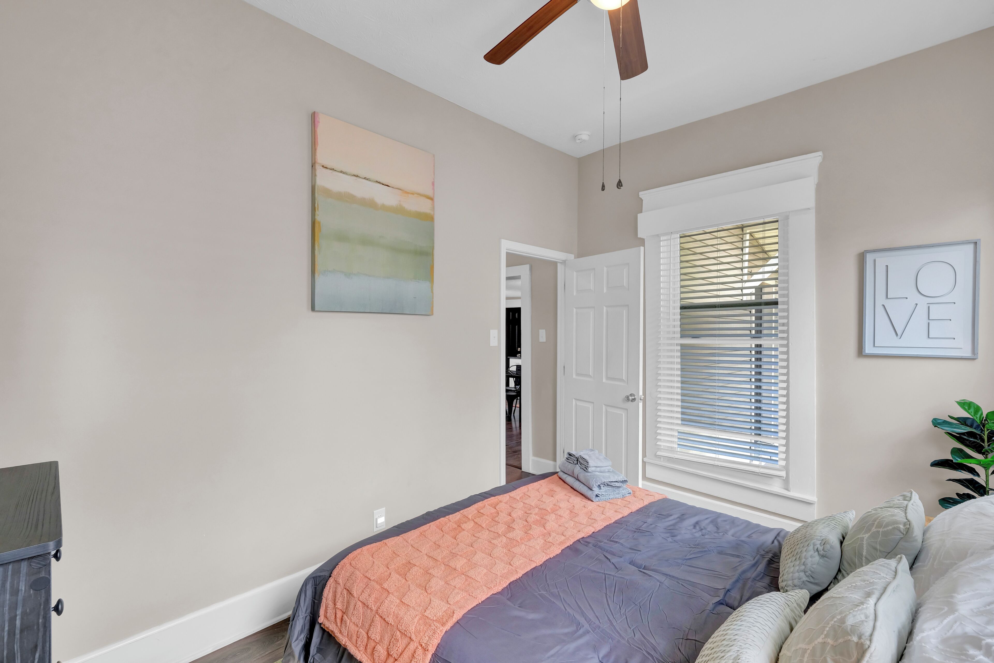 A bedroom with a bed, a ceiling fan, a window with blinds, a door, and a wall with a painting.