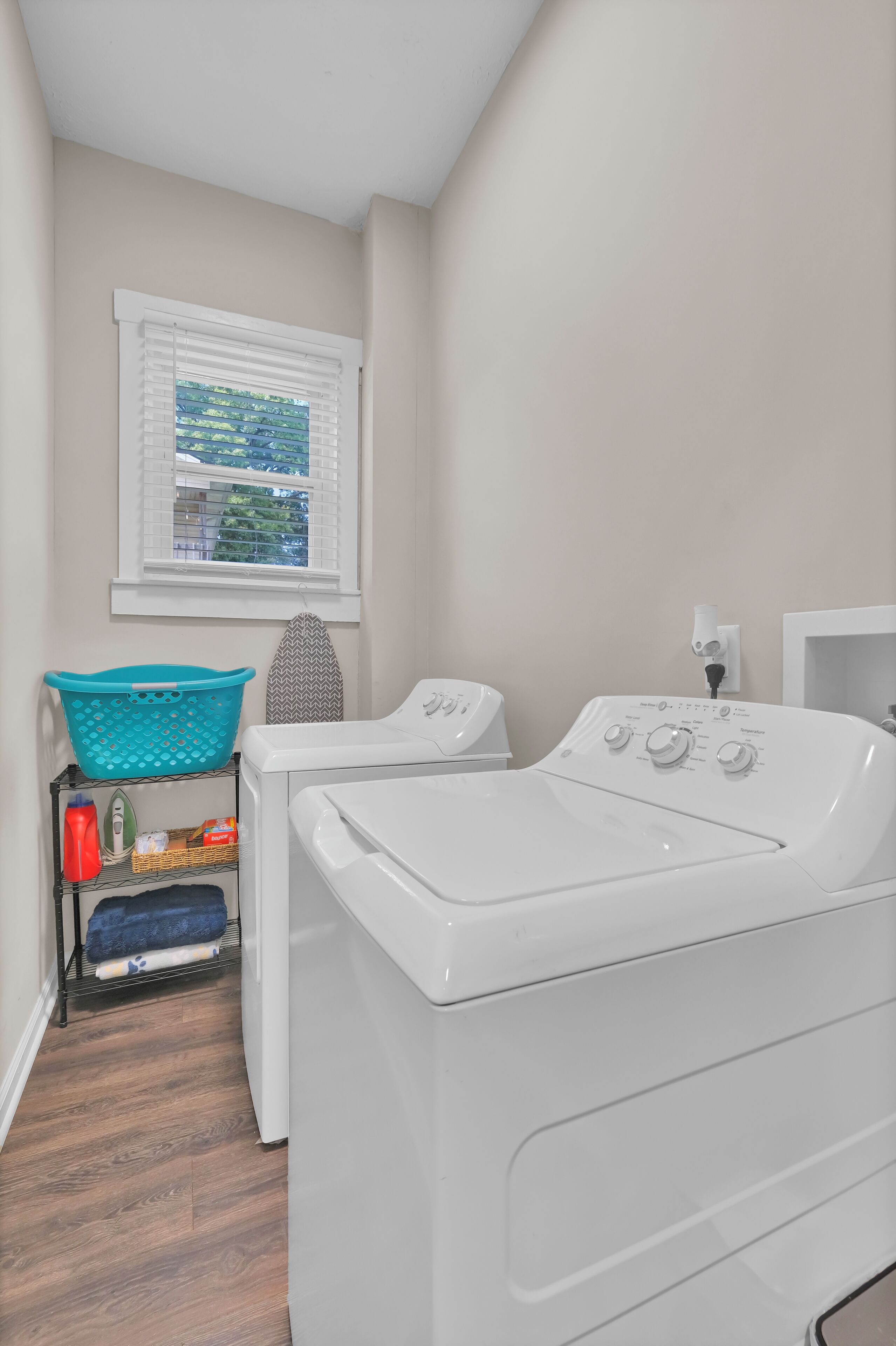 A laundry room with two front-loading washers and dryers, a window with blinds, a laundry basket, and a shelf with cleaning supplies.