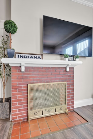 A fireplace with a brick wall, a mounted TV above it, and a sign that reads 'INDIANA' on the mantel. - Sleeps 8 • Fenced Yard and Firepit • 8 Minutes to Louisville (Jeffersonville)