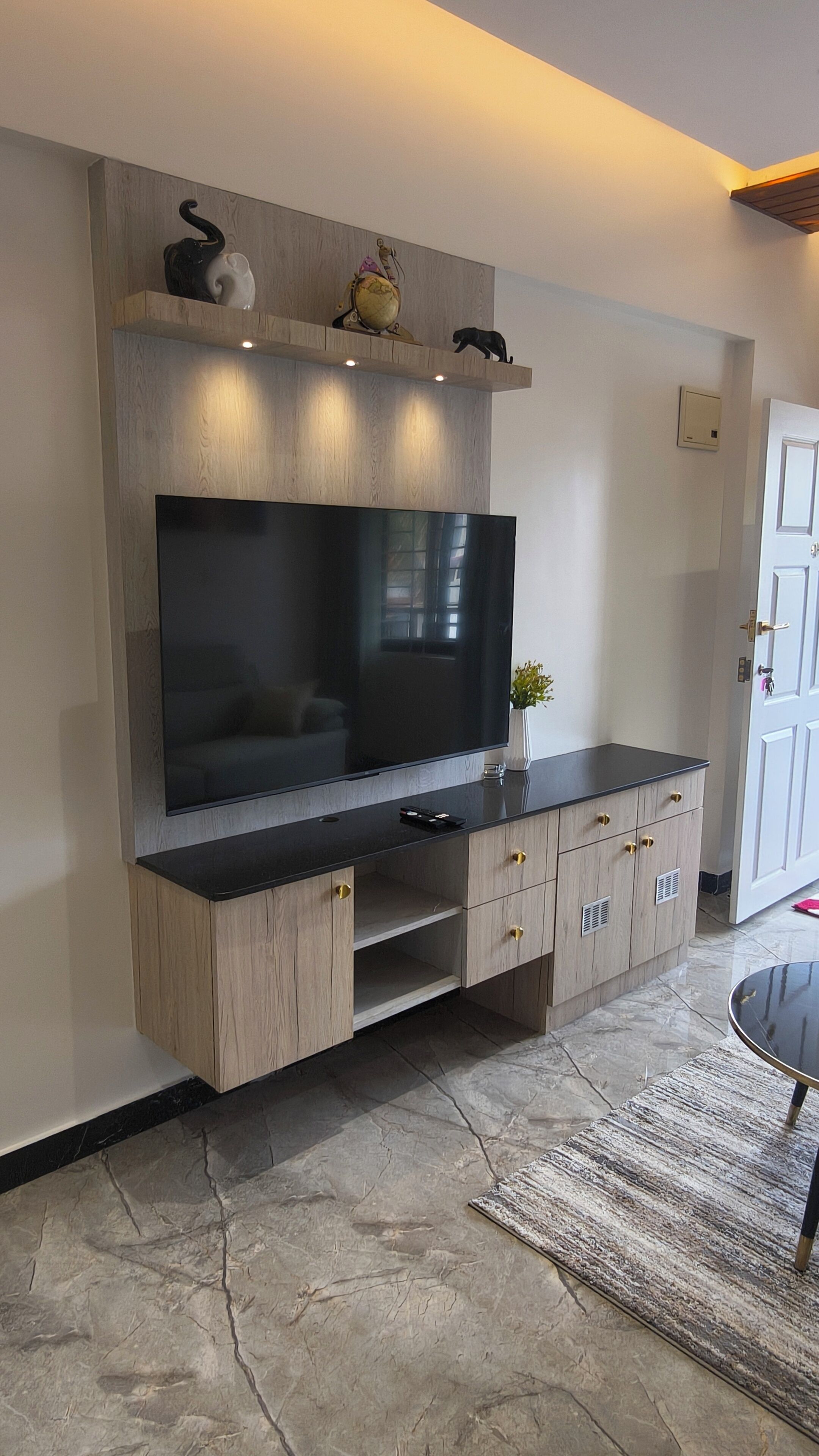 A modern living room with a large flat-screen TV mounted on a wooden wall unit, a small plant on the unit, and a door to the right.