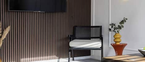 Una sala de estar moderna con un televisor de pantalla plana montado en la pared, una silla negra con cojín blanco, un revestimiento de pared de listones de madera, una mesita naranja pequeña y una planta en maceta con follaje amarillo y verde.