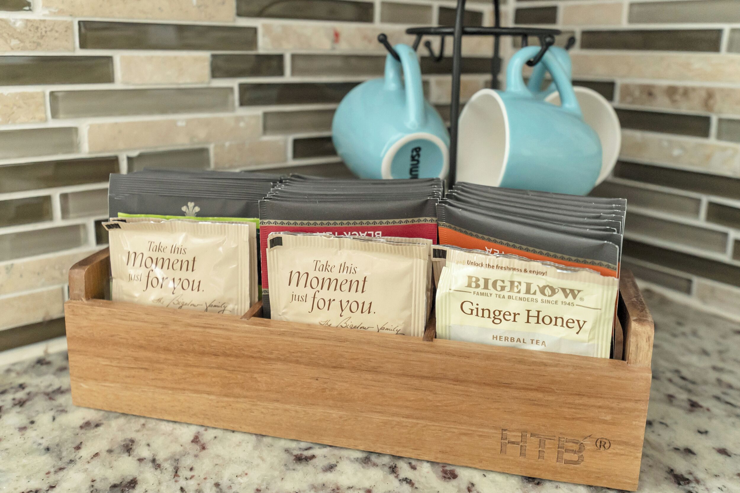 A wooden tray holding tea bags with messages, placed on a kitchen counter with a tiled backsplash and blue kitchen utensils in the background.