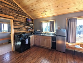 Cocina con paredes de madera, estufa, armarios, refrigerador y ventana con cortinas.