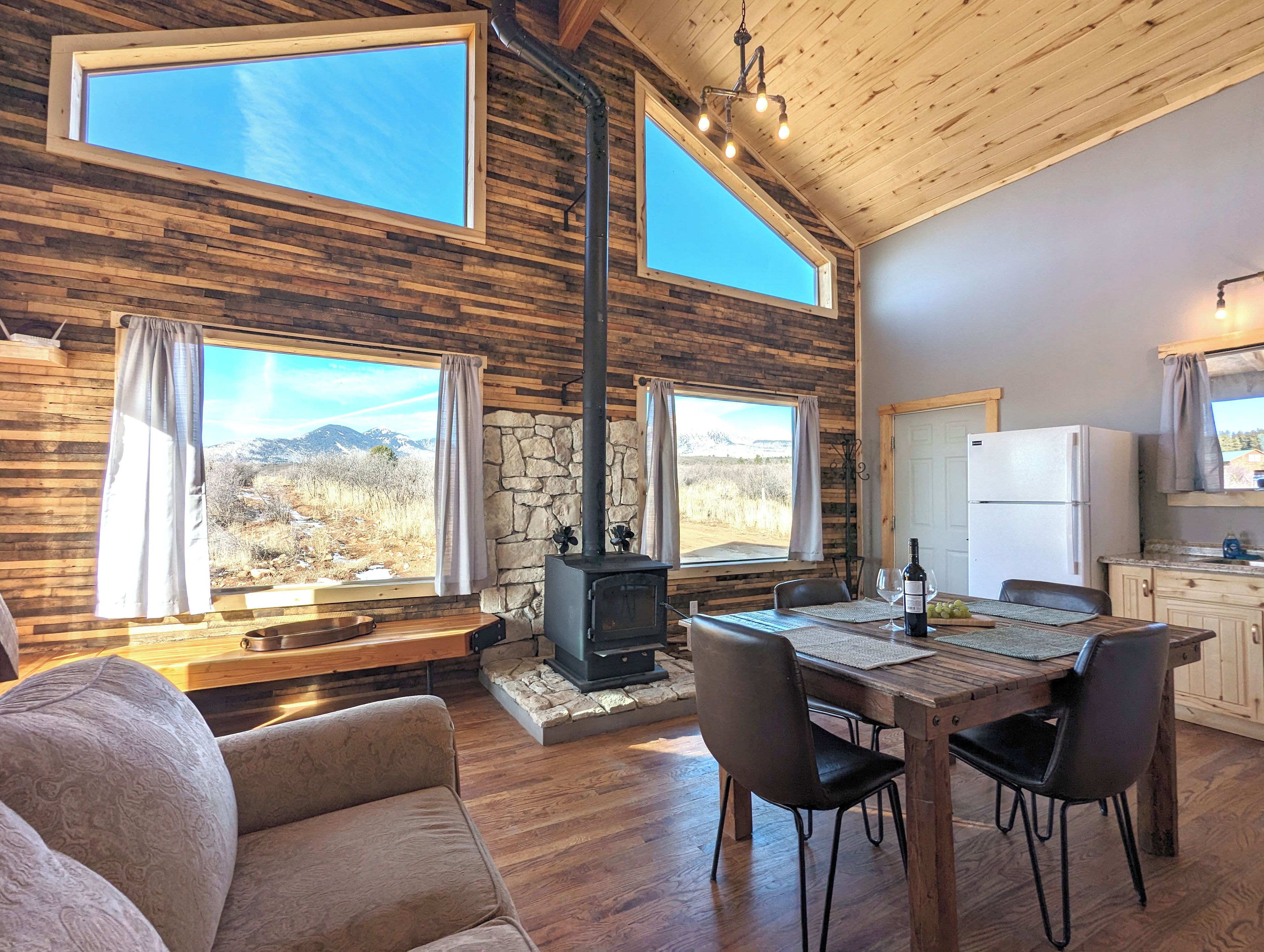 Interior acogedor de una cabaña con chimenea de piedra, mesa de comedor de madera y ventanas amplias con vistas a un paisaje montañoso.
