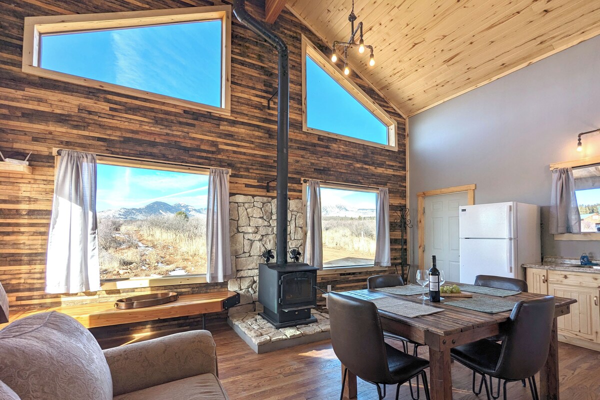 A cozy cabin interior with a stone fireplace, wooden dining table, and large windows overlooking a mountainous landscape.