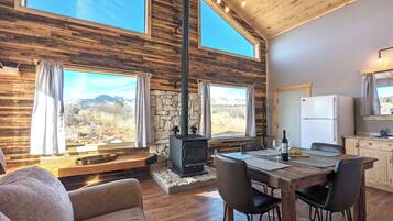 Interior acogedor de una cabaña con chimenea de piedra, mesa de comedor de madera y ventanas amplias con vistas a un paisaje montañoso.