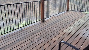 Una terraza de madera con barandilla metálica, una única silla de metal y vista a un paisaje boscoso.