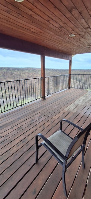 Um deck de madeira com guarda-corpo de metal, uma única cadeira de metal e vista para uma paisagem arborizada.