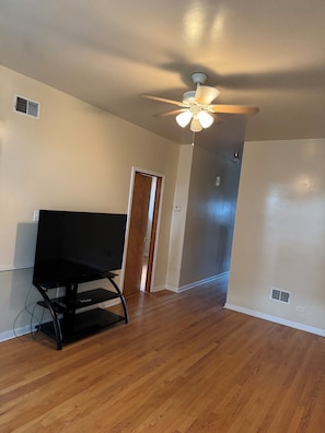 A room with a ceiling fan, a flat-screen TV on a stand, wooden flooring, and a door leading to another room. - Welcoming 2-bedroom building with AC in vibrant Chicago (Chicago)