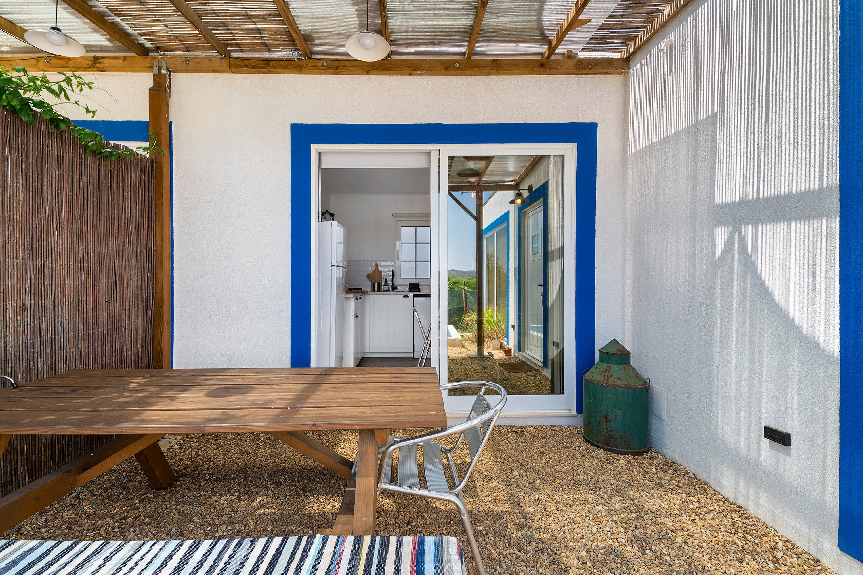 Un espacio al aire libre cubierto con una mesa de madera y una silla de metal, una cocina visible a través de un arco abierto y una puerta con marco azul.