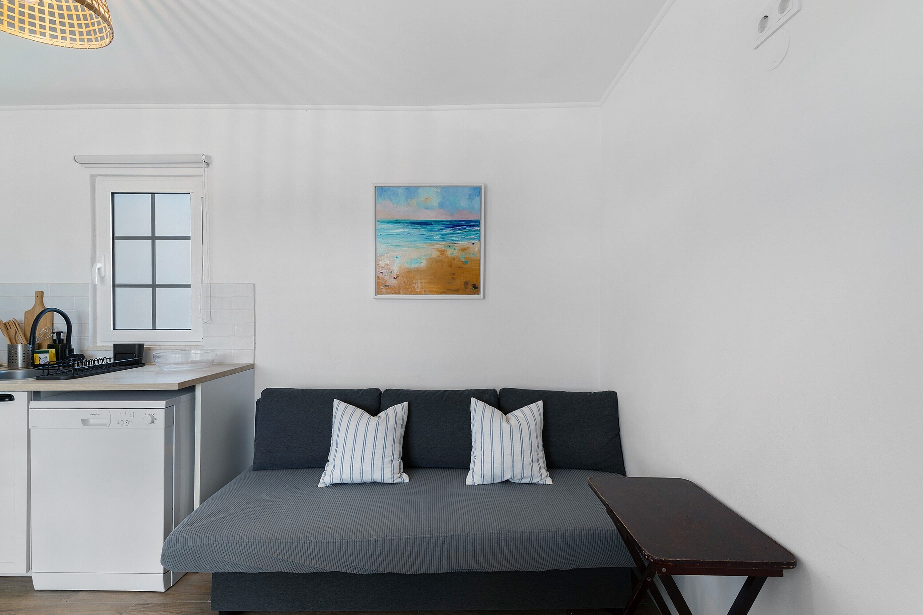 Un espacio habitable compacto con kitchenette, un sofá con cojines a rayas y un cuadro con motivos playeros en la pared.