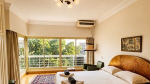 A hotel room with a large bed, a view of greenery and buildings through the window, a ceiling fan, and a painting on the wall. - Royal Retreat in Dokki • Spacious with Nile View (Giza Governorate)