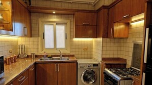 A kitchen with wooden cabinets, a washing machine, a stove, and a window with blinds. - Royal Retreat in Dokki • Spacious with Nile View (Giza Governorate)