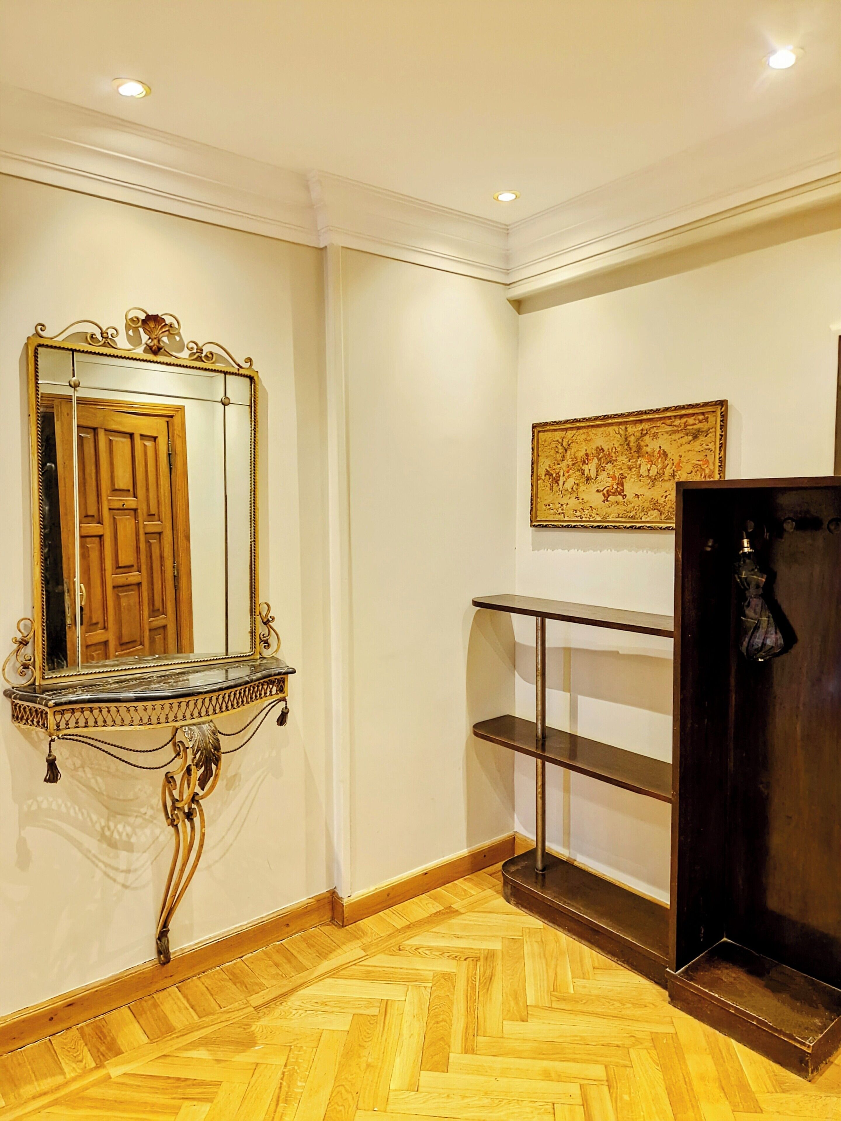 Ein Zimmer mit Holztür, einem Spiegel mit aufwendigem Metallrahmen, Holzboden und einem an der Wand montierten Regal.
