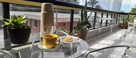 Un balcón con una mesa de vidrio, una cafetera, una taza amarilla, un plato con un pastelito y plantas en macetas.