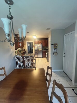 Zona pranzo con tavolo e sedie in legno, un lampadario e una cucina sullo sfondo.