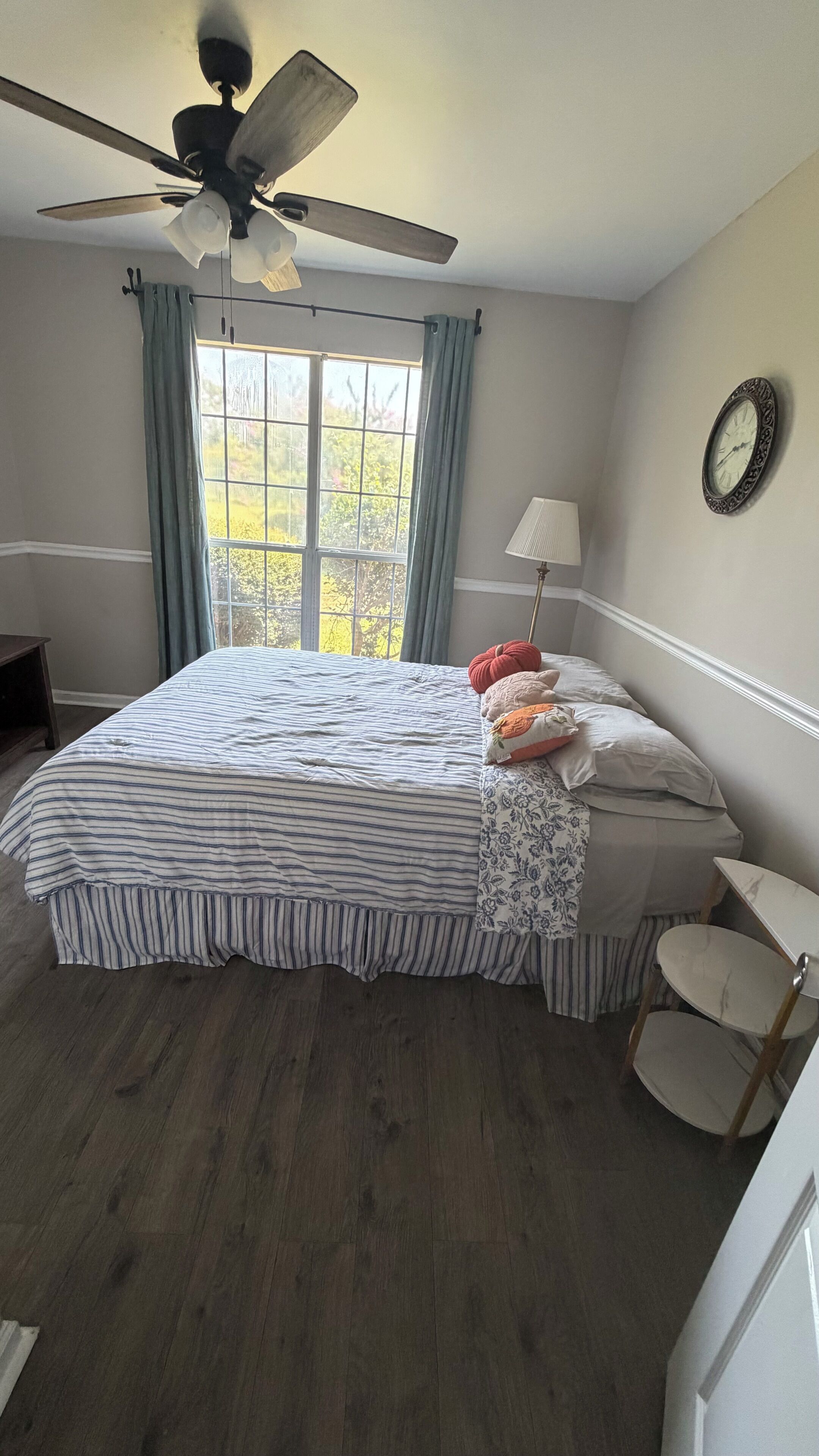 A bedroom with a bed, a ceiling fan, a window with curtains, a bedside table, and a wall clock.