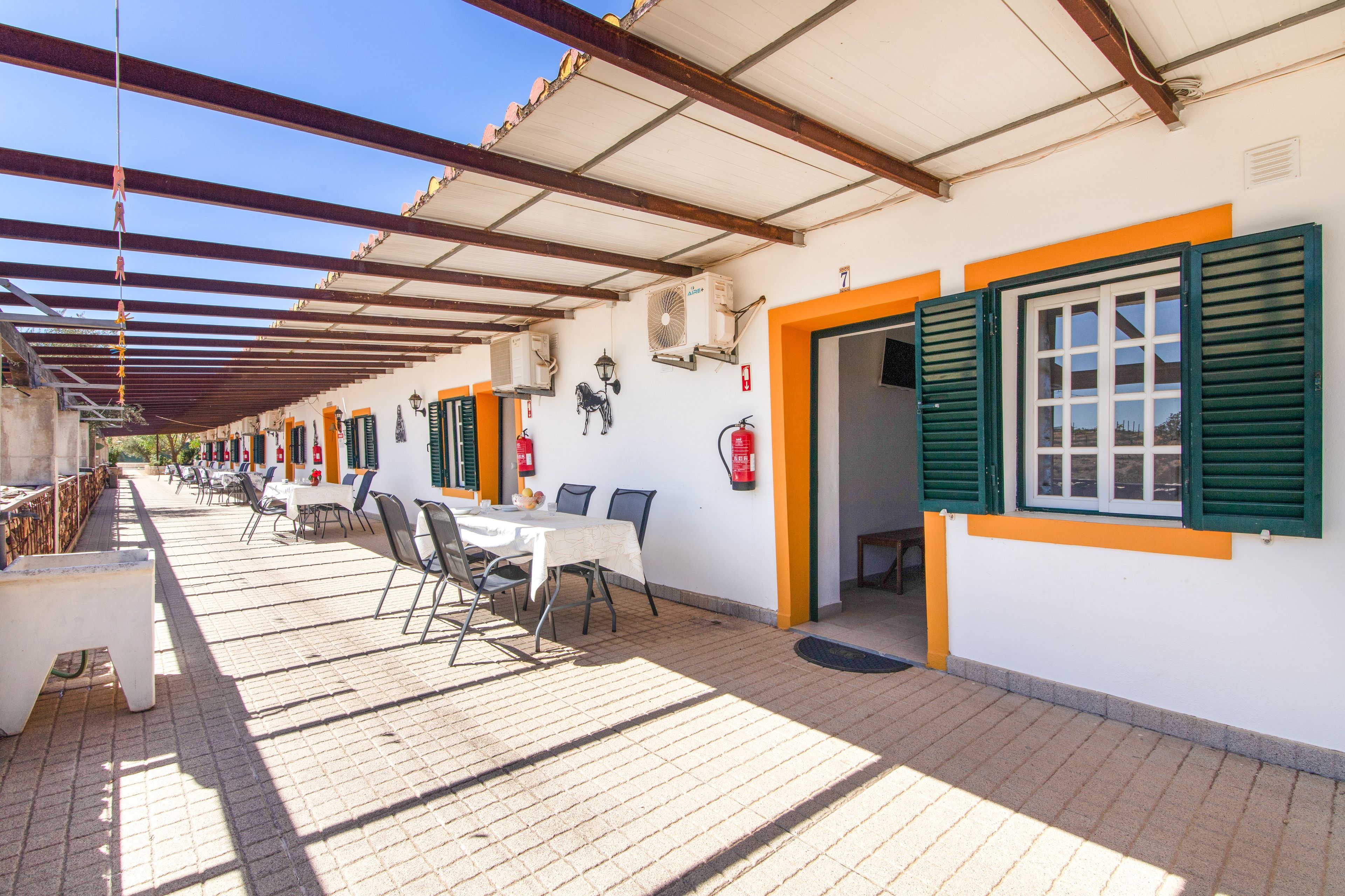 Een overdekt terras buiten, met tafels en stoelen, een brandblusser en een aan de muur bevestigde airconditioning.