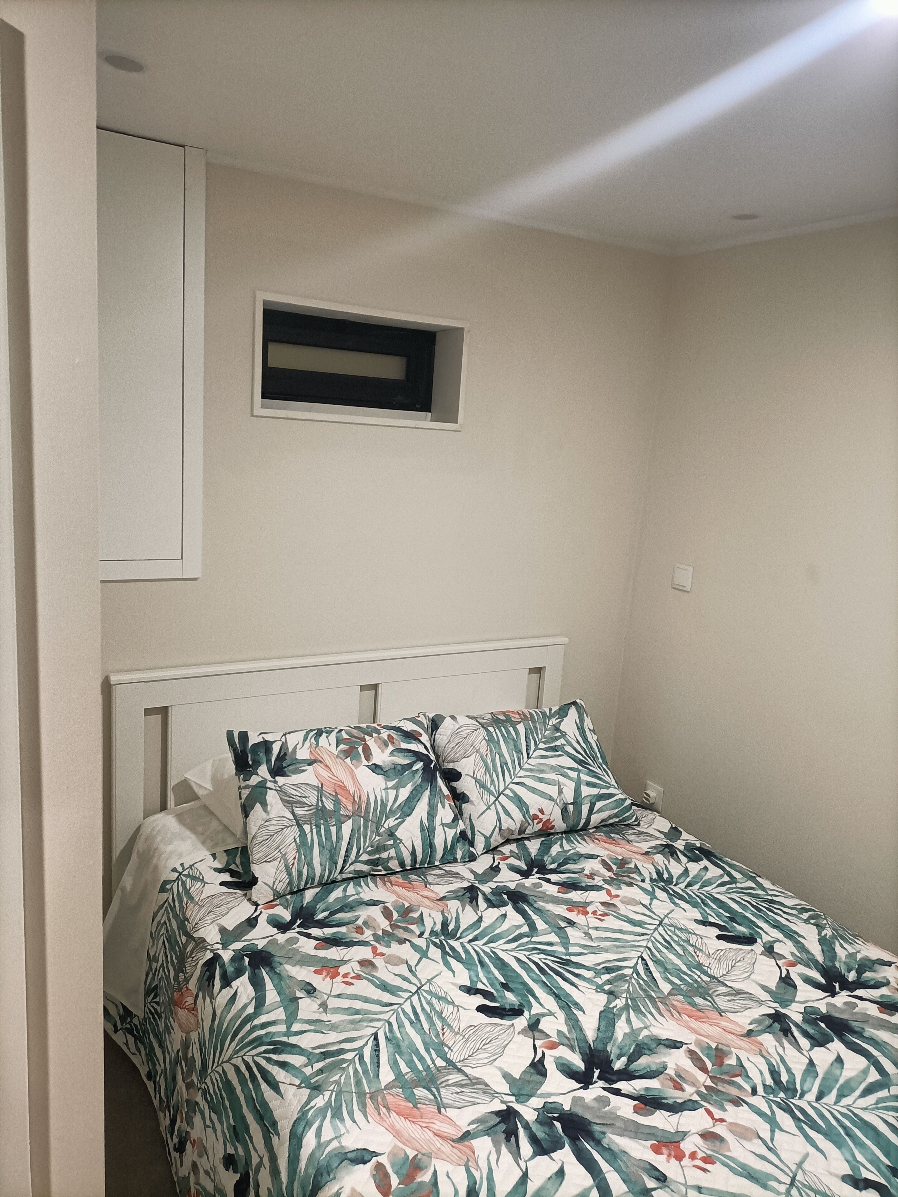 A bed with a floral bedspread, a white headboard, and a small window above the bed.