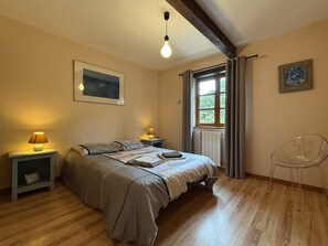 Ein Schlafzimmer mit einem Bett, Nachttischlampen, einem Fenster mit Vorhängen, einem Heizkörper, einem transparenten Stuhl und einem gerahmten Bild an der Wand.