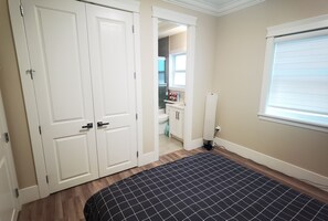 Een slaapkamer met een bed, een kledingkast, een zichtbare badkamer door een geopende deur en een raam met zonwering.