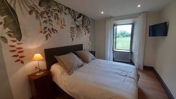 Et soveværelse med seng, natborde, lampe, tv ophængt på væggen og et vindue med udsigt til grønne områder.
