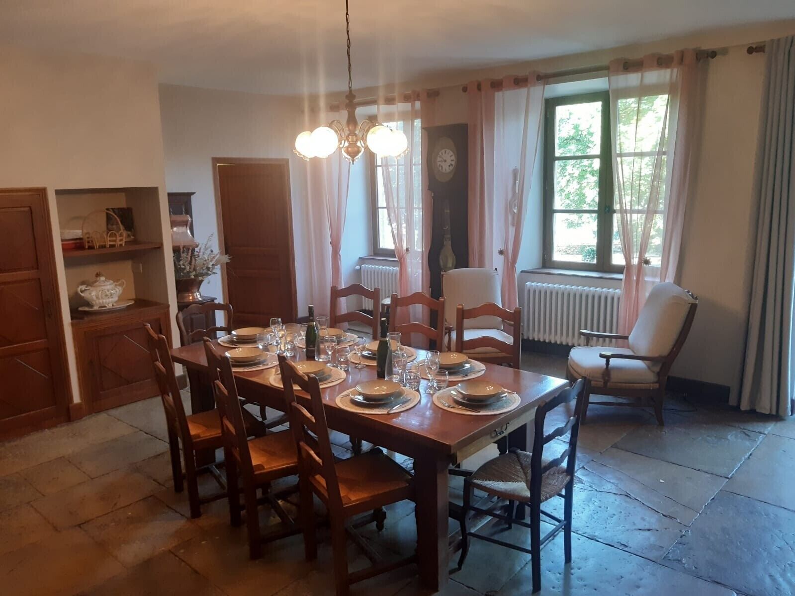 Een eetkamer met een houten tafel voor zes personen, omringd door stoelen, een kroonluchter en een uitzicht op het landschap door een raam met gordijnen.