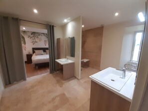 Quarto de hotel com cama, banheiro com pia e espelho, e chuveiro.