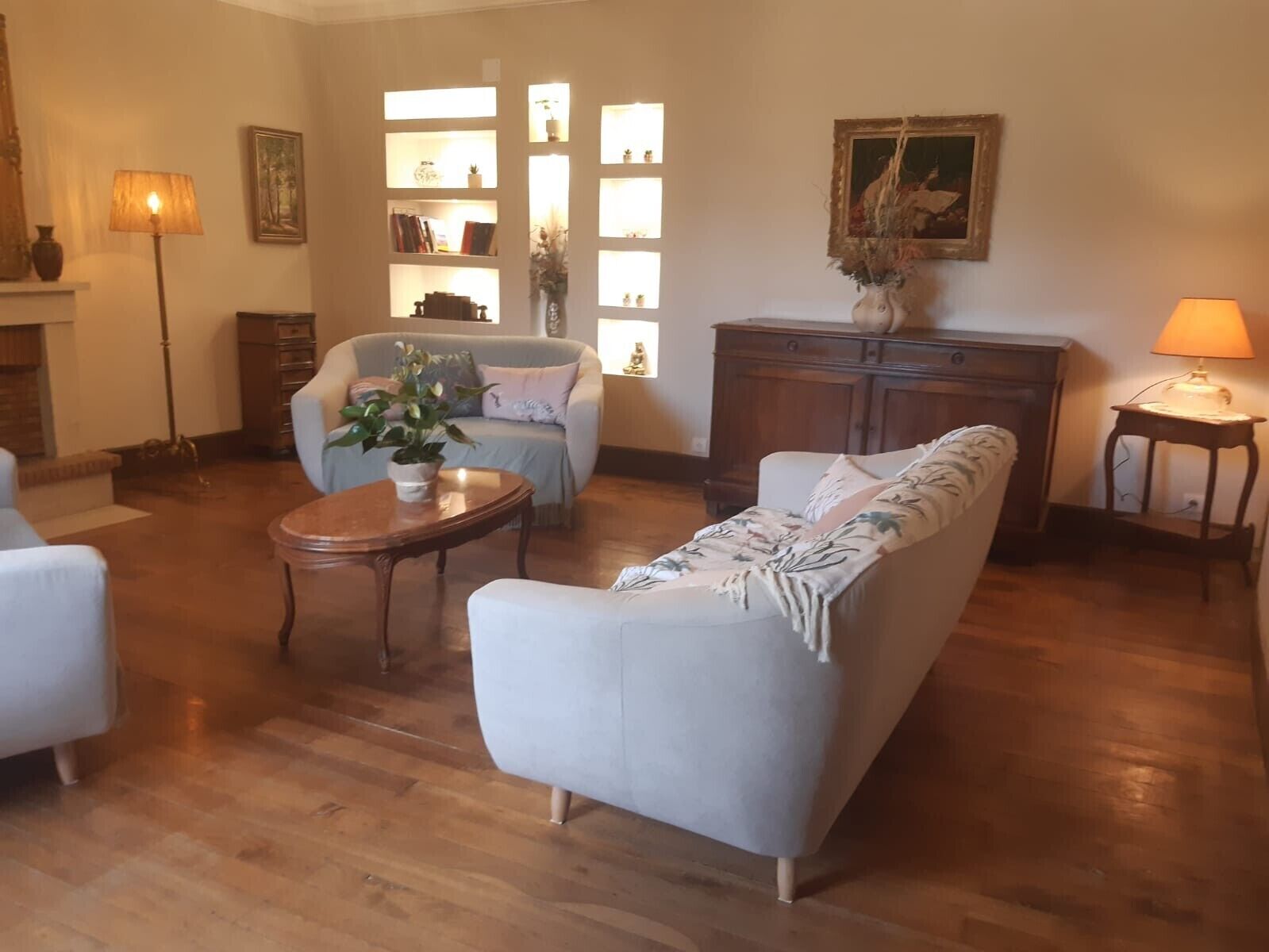 Un salon comprenant un canapé, un fauteuil et une petite table sur laquelle est posée une plante. On y trouve également une cheminée, une lampe et un meuble contenant des objets décoratifs.