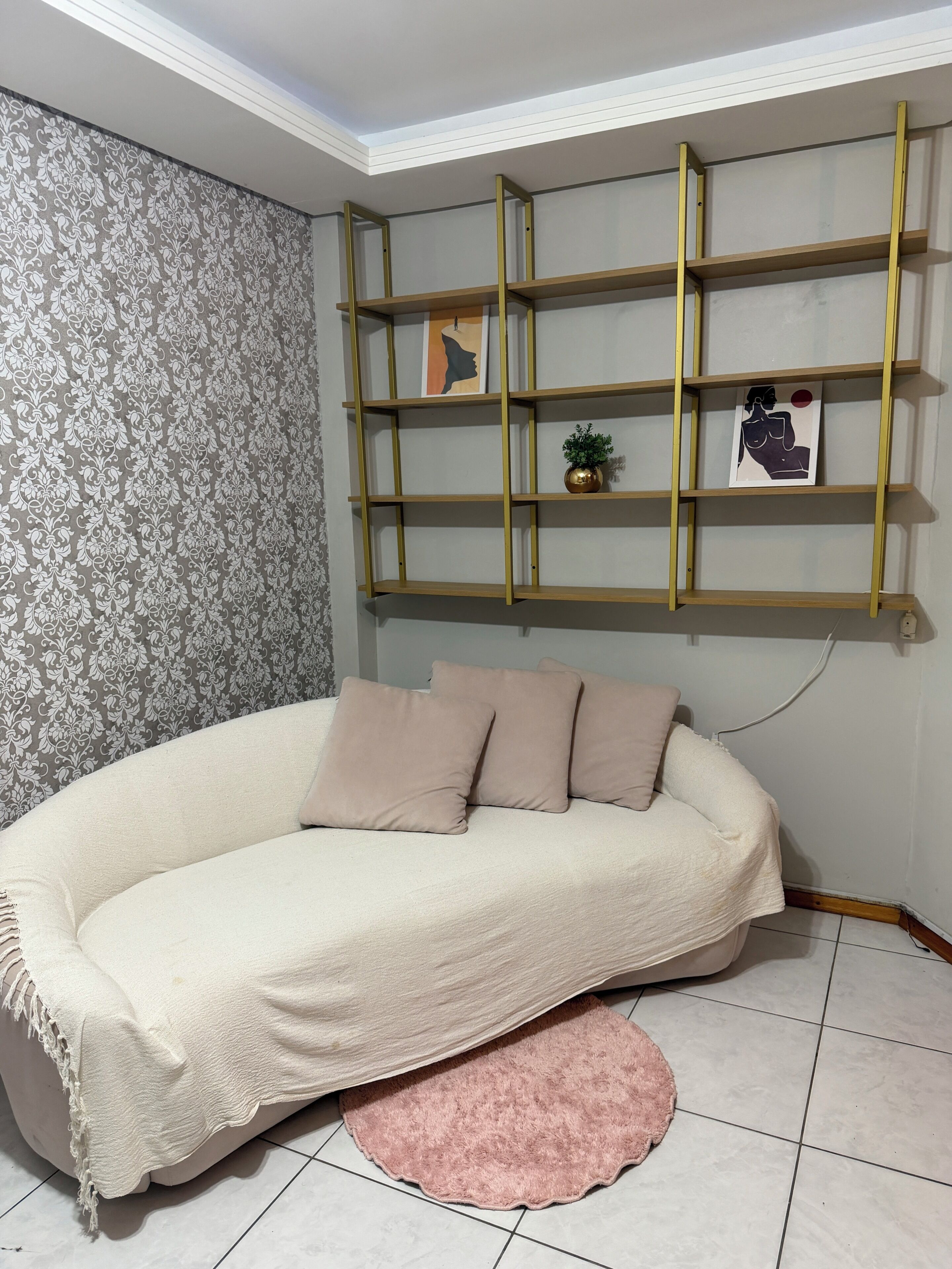 Ein Zimmer mit strukturierter Tapete, einem beigen Sofa mit Decke, einem rosafarbenen Teppich und einem an der Wand befestigten Regal mit Kunstwerk und einer kleinen Pflanze.