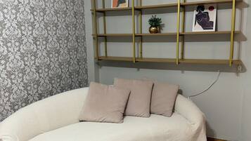 Ein Zimmer mit strukturierter Tapete, einem beigen Sofa mit Decke, einem rosafarbenen Teppich und einem an der Wand befestigten Regal mit Kunstwerk und einer kleinen Pflanze.